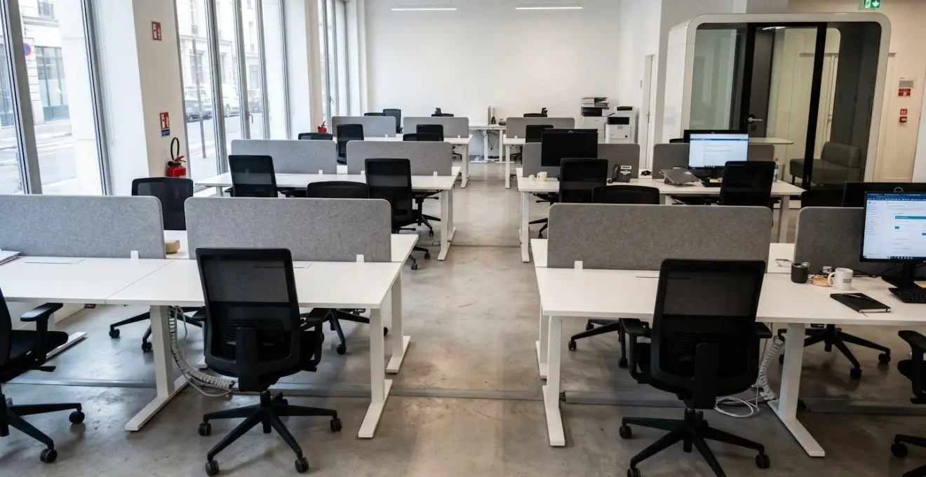 Open space contemporain vide avec panneaux acoustiques gris fixés sur bureaux blancs, fauteuils ergonomiques noirs à dossier haut et cabine insonorisée vitrée en arrière-plan, baigné de lumière naturelle par grandes fenêtres