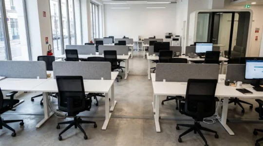Open space contemporain vide avec panneaux acoustiques gris fixés sur bureaux blancs, fauteuils ergonomiques noirs à dossier haut et cabine insonorisée vitrée en arrière-plan, baigné de lumière naturelle par grandes fenêtres